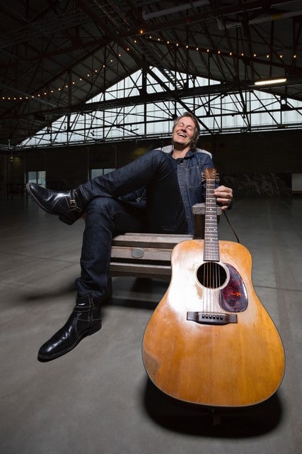 Jim Cuddy sitting back in a wooden chair and holding a guitar